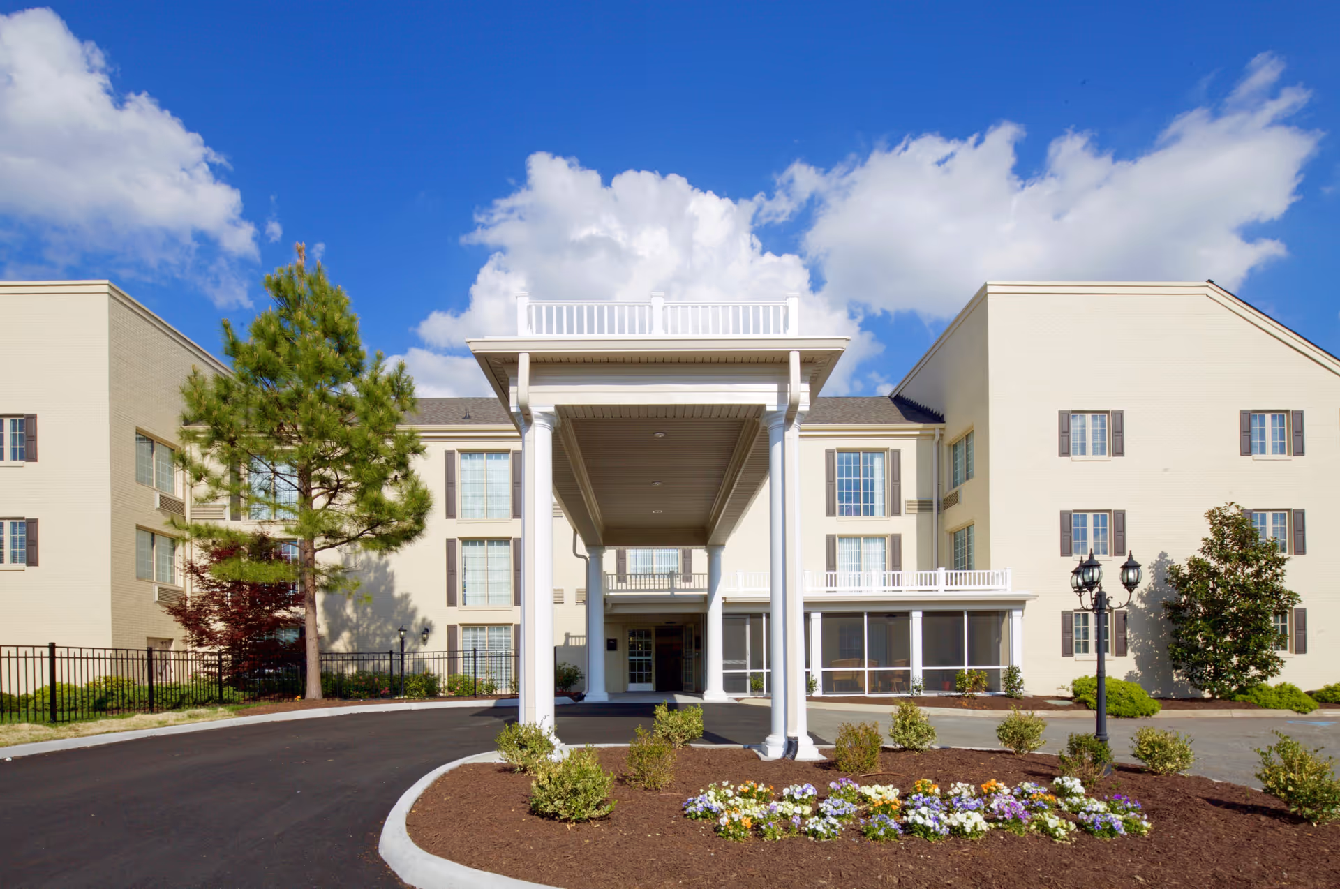 Entrance of a multi-story beige senior living building with a covered porte-cochere, landscaped flower beds, and a blue sky.