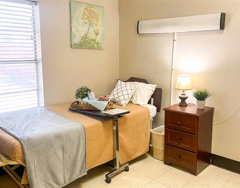 A small resident bedroom with a single bed dressed in beige and gray linens, an overbed table, and a wooden nightstand holding a lamp and plant.