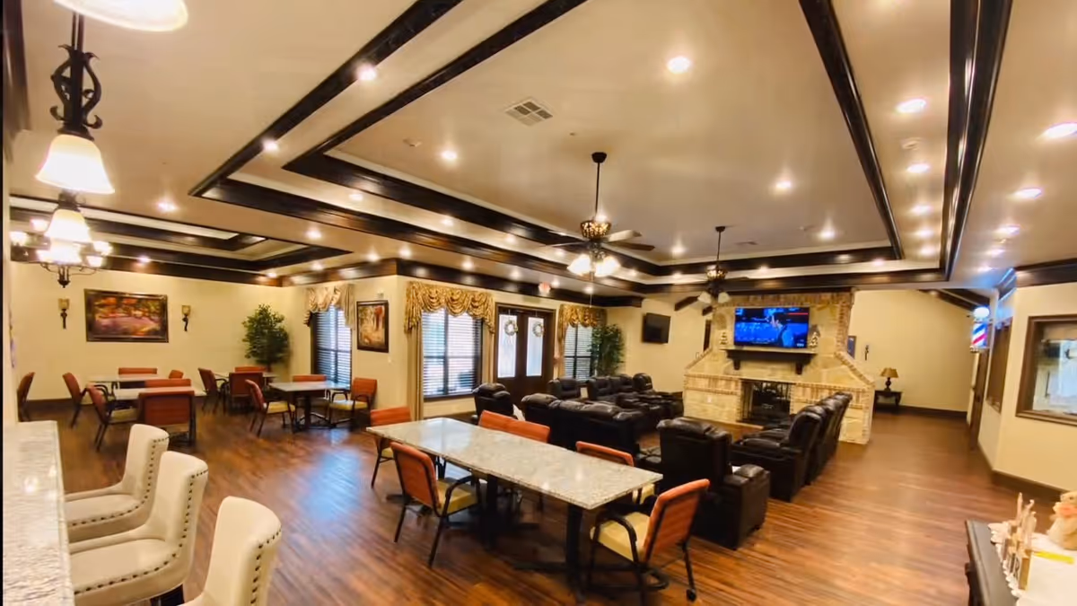 A spacious and well-lit common area in Avalon Memory Care featuring multiple seating arrangements including tables with chairs and a cluster of black leather recliners facing a large flat-screen TV mounted above a stone fireplace. The room has wooden floors, decorative ceiling beams with recessed lighting, ceiling fans, and large windows with curtains. There are paintings and plants decorating the walls.