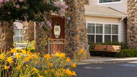Stone front entrance of a senior living facility with flowering garden and bench seating.