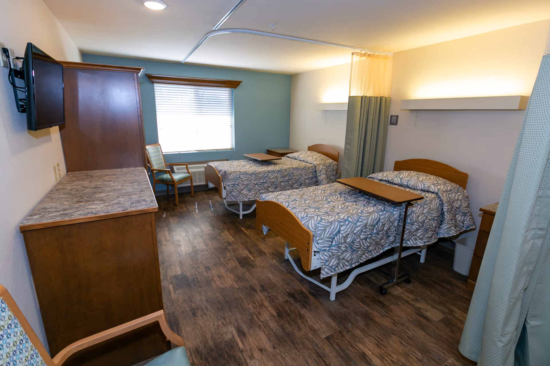 A shared patient room with two hospital-style beds, overbed tables, chairs, a dresser, and a window.