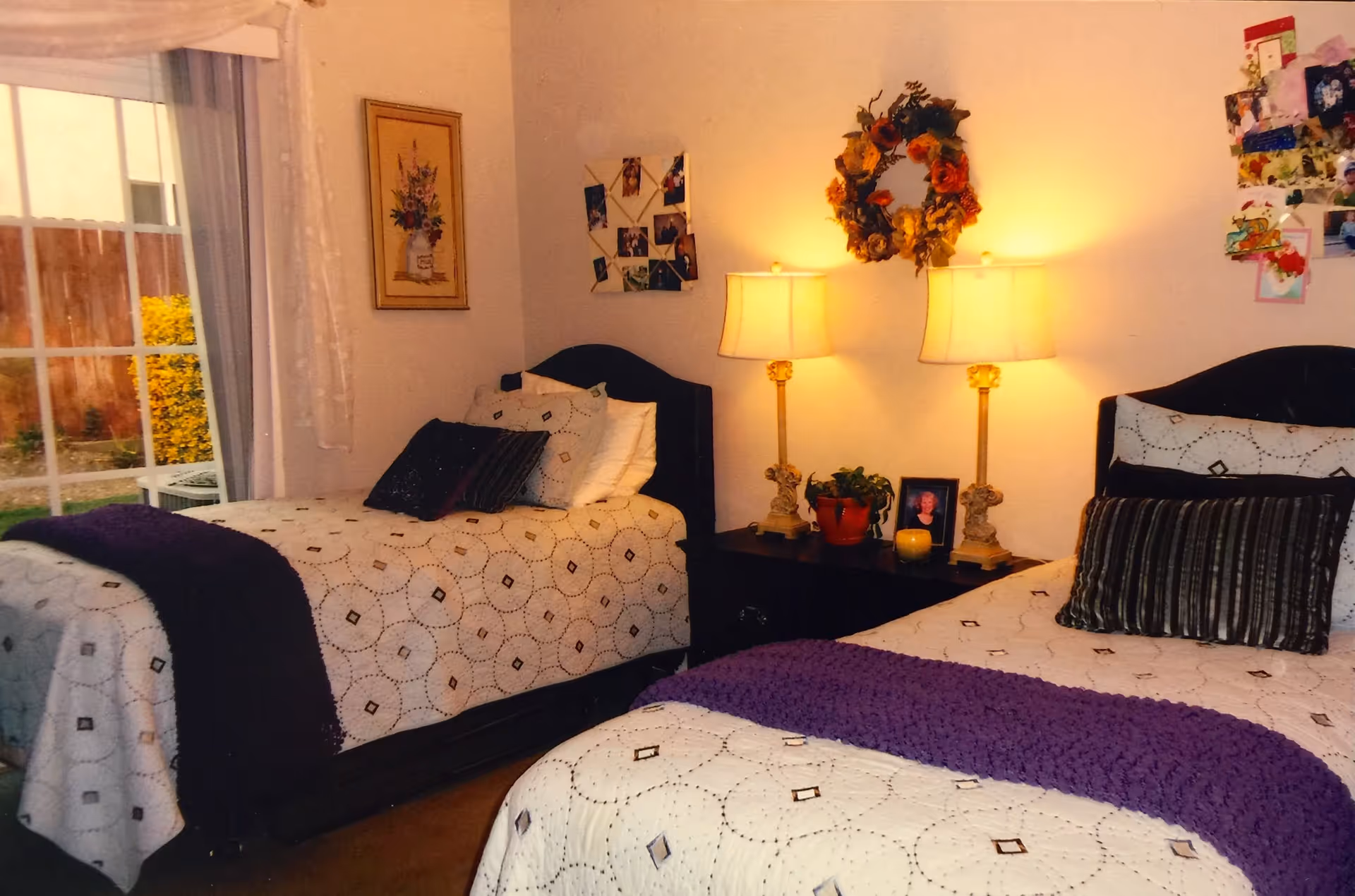 Two twin beds with patterned quilts and purple throws flank a nightstand with twin lamps and wall decor beside a sliding glass door.