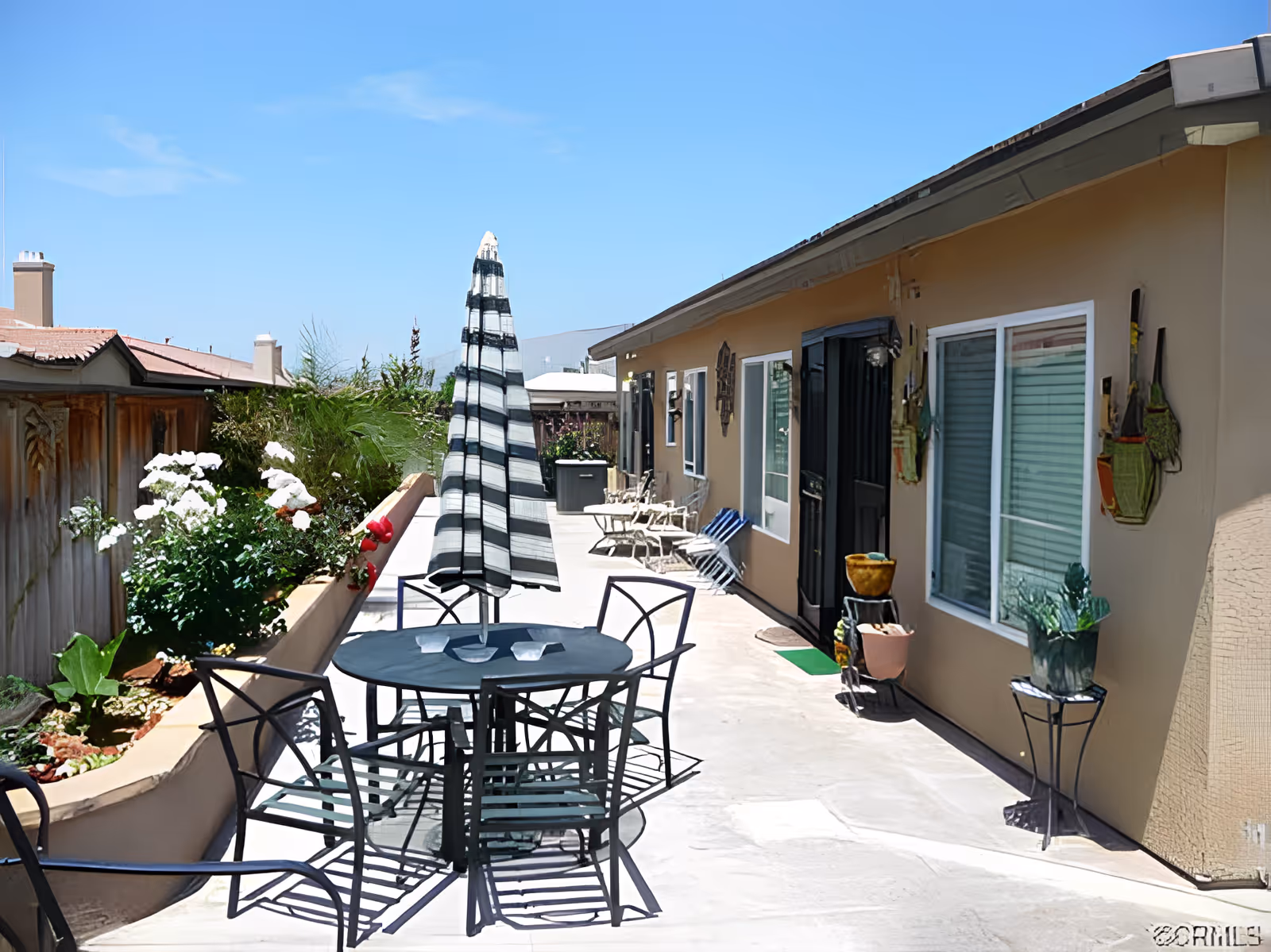 Outdoor patio walkway with a round table, striped umbrella, chairs, potted plants, and adjacent building doors and windows.