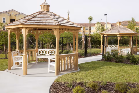 Two wooden gazebos with shingled roofs and white benches inside, situated on a paved pathway surrounded by green grass and landscaped plants, with residential buildings and palm trees in the background.