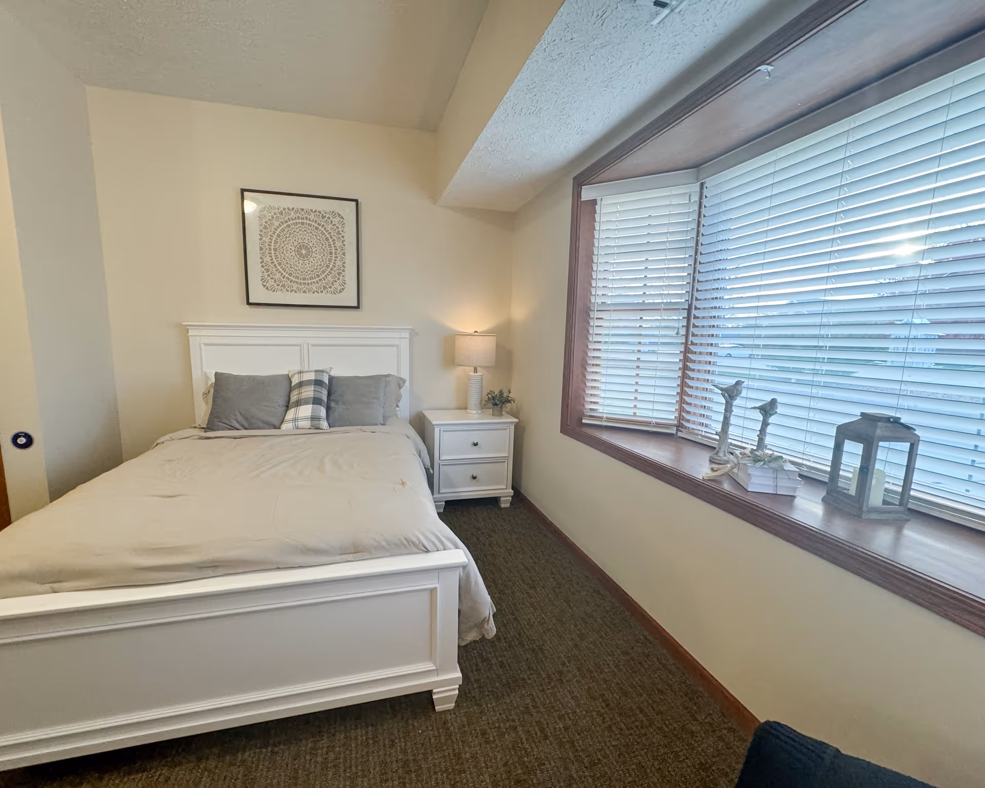 A cozy bedroom with a white wooden bed frame and beige bedding. There are three pillows on the bed, two gray and one plaid. A white nightstand with two drawers is beside the bed, holding a lamp and a small plant. A large window with white blinds lets in natural light, and the windowsill is decorated with two bird figurines, a small stack of books, and a lantern. A framed decorative artwork hangs on the wall above the bed.