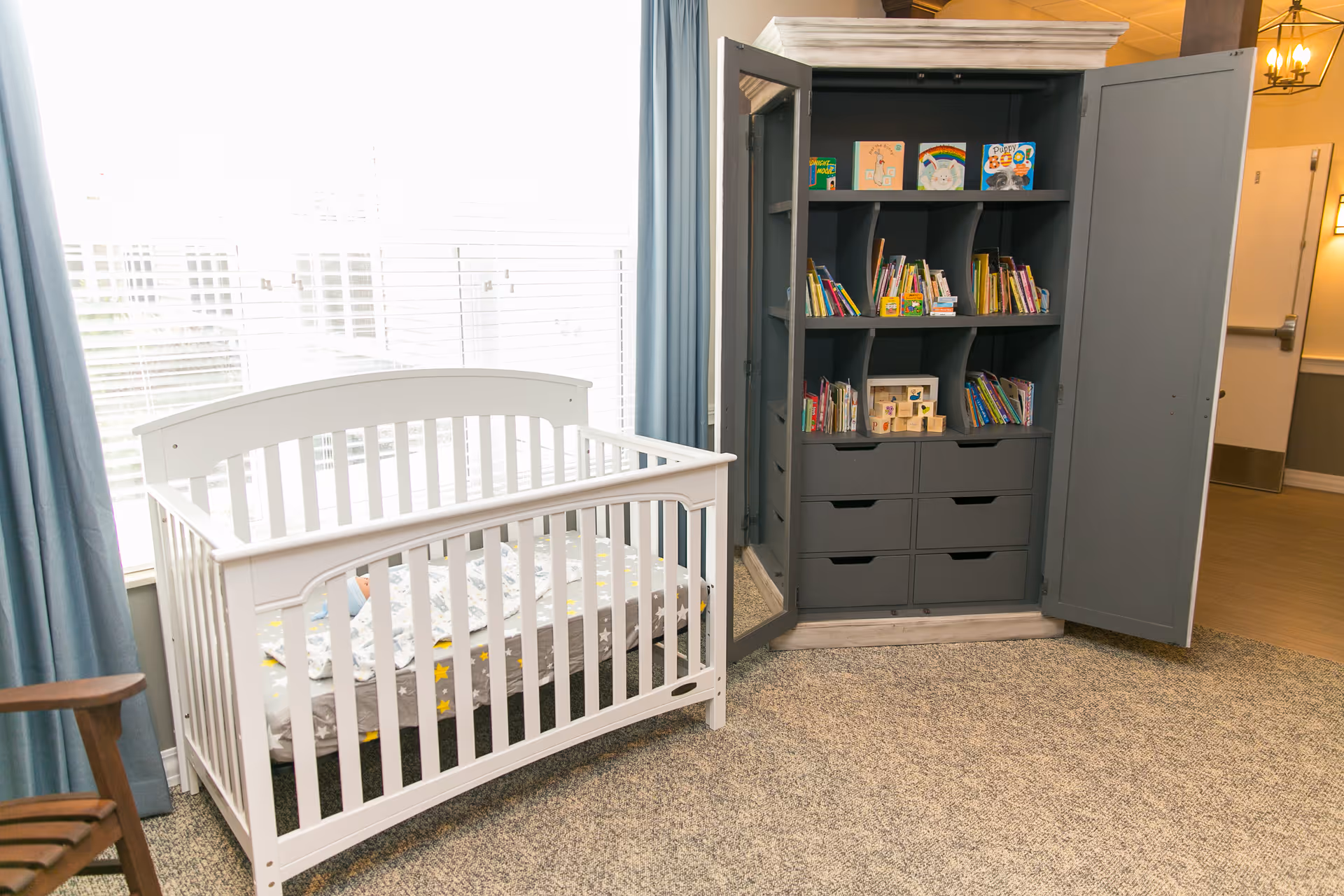 A bright room with a white baby crib near a large window with blue curtains. Next to the crib is a gray cabinet with open doors, filled with children's books and toys. The room has carpeted flooring and a wooden chair partially visible on the left.