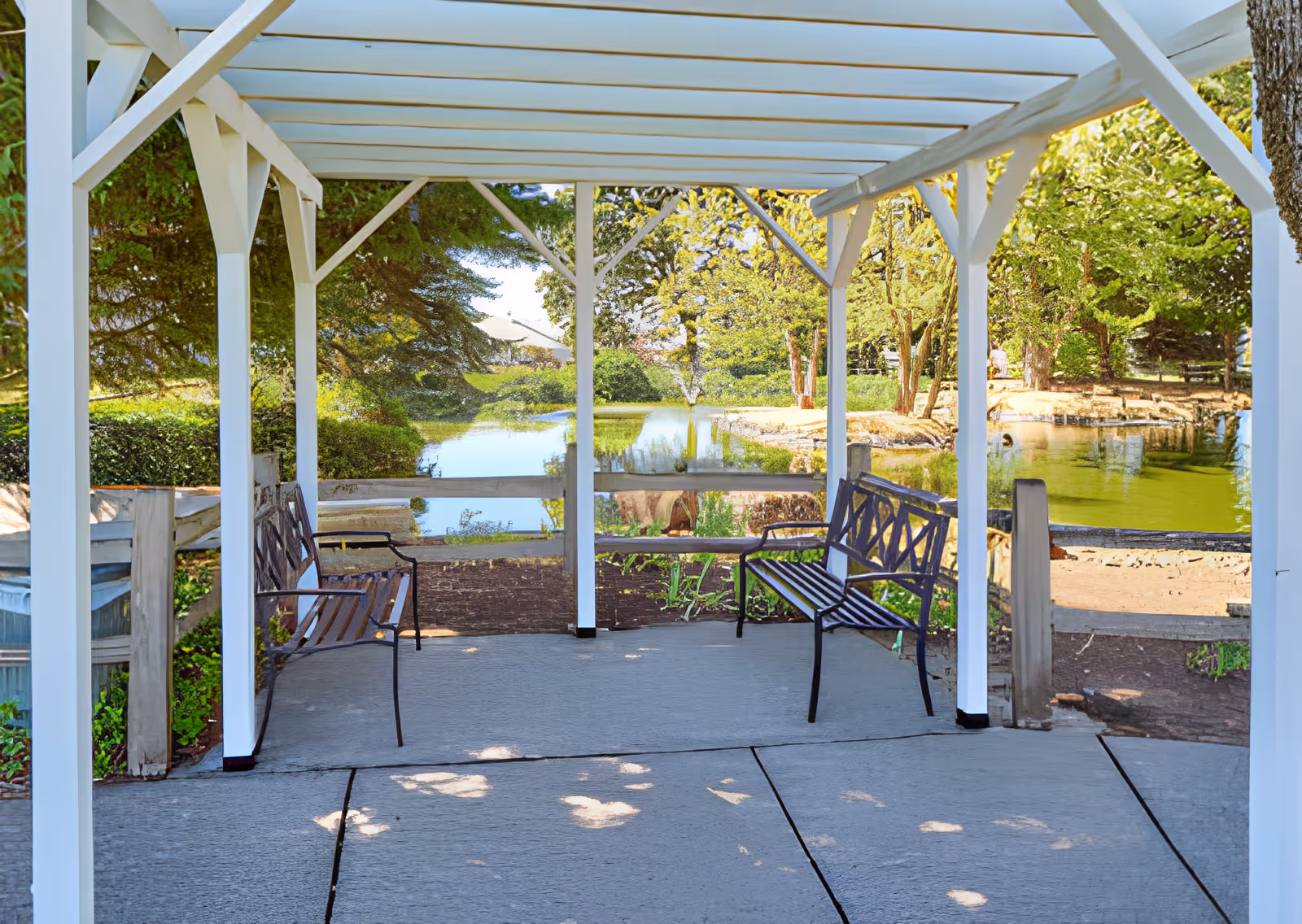 A white pergola with two black metal benches facing each other, overlooking a serene pond surrounded by lush green trees and bushes on a sunny day.