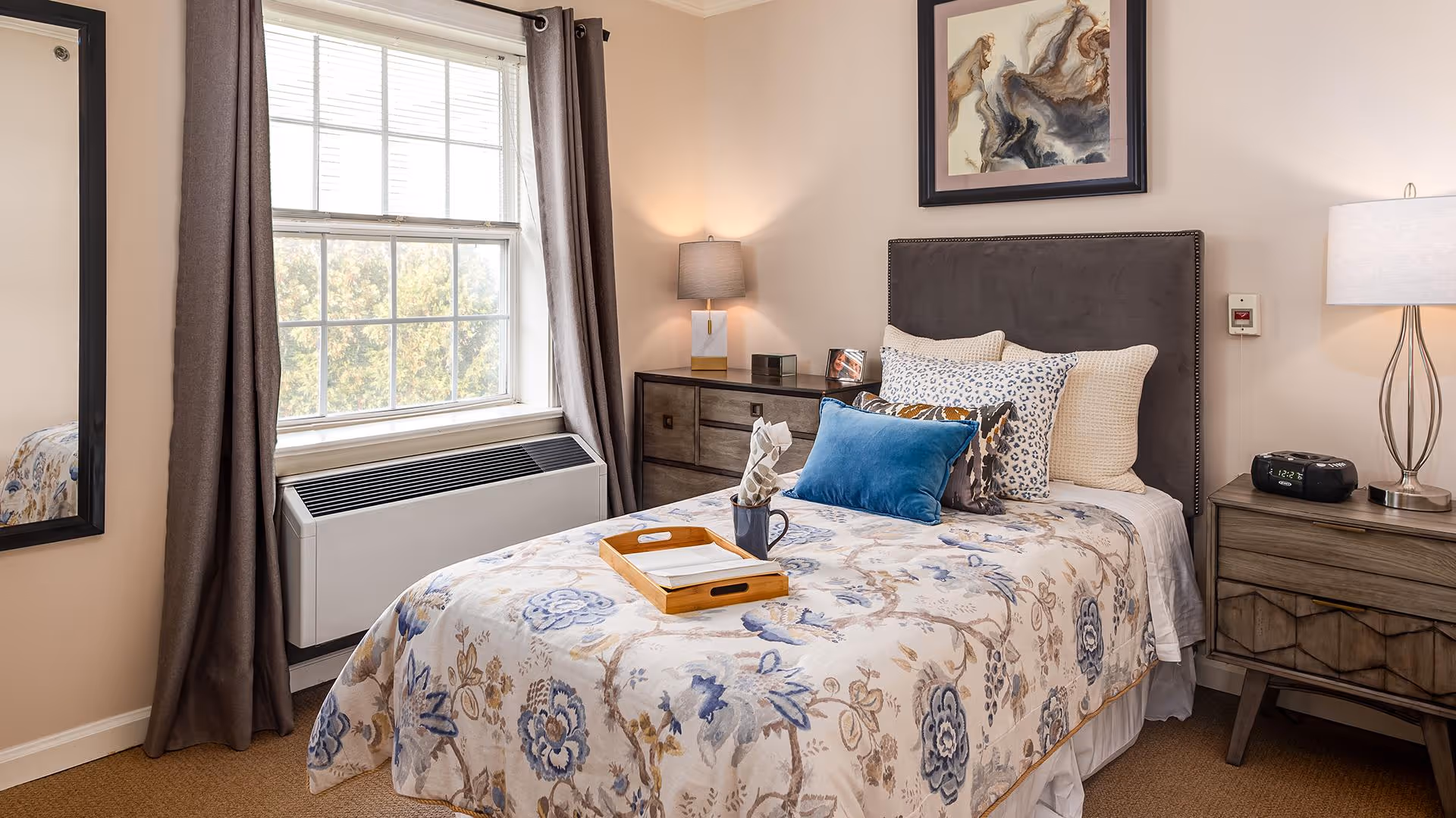 A cozy bedroom with a single bed covered in a floral patterned comforter and multiple pillows, including a blue accent pillow. There is a wooden tray with a cup and napkin on the bed. The room has a large window with gray curtains, a dresser with a lamp and picture frame, a nightstand with a lamp and digital clock, and a framed abstract painting on the wall above the bed.