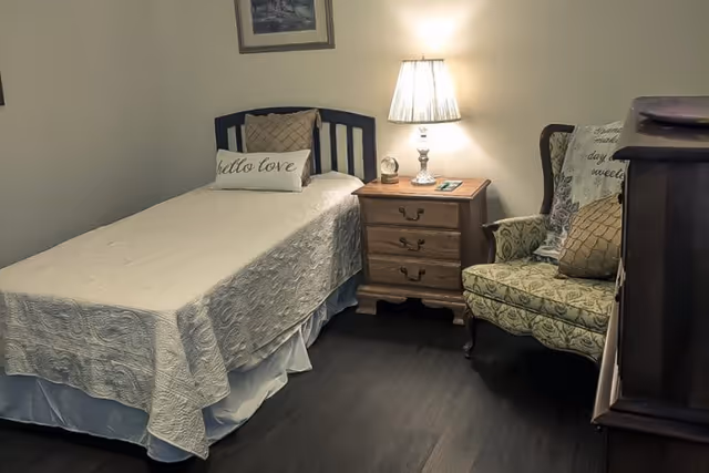 A cozy bedroom containing a single bed with decorative pillows, a wooden nightstand with a lamp, and an upholstered armchair.