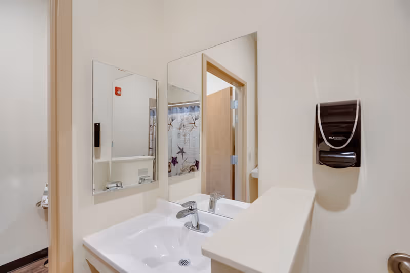 A clean bathroom sink area with a white countertop and a chrome faucet. Two mirrors are mounted on the wall above the sink, reflecting a partially open door and a shower curtain with starfish and seashell designs. A black soap dispenser is mounted on the wall to the right of the sink.