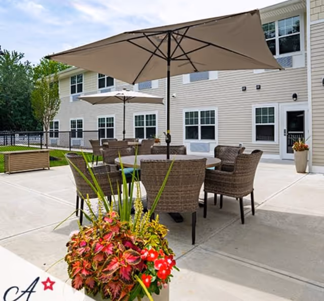 Outdoor patio area at All American Assisted Living at Tinton Falls featuring round tables with wicker chairs and large beige umbrellas. There is a planter with colorful flowers in the foreground and a two-story building with multiple windows in the background.