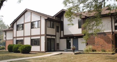 Exterior view of a two-story residential building with a combination of brick and light-colored panel siding, surrounded by grass and some bushes, with a tree partially visible on the right side.