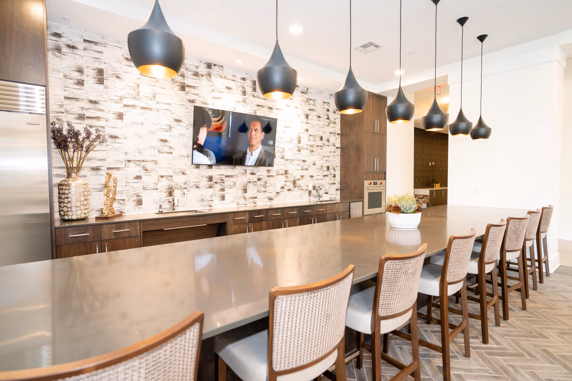 A modern communal kitchen/dining area featuring a long island with bar stools, pendant lights, a wall-mounted TV, and wood cabinetry.