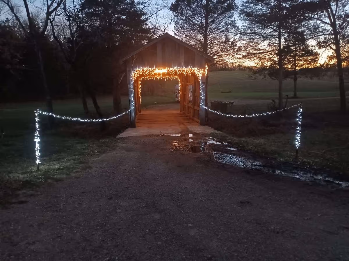 A small wooden covered bridge decorated with white string lights at dusk, with illuminated posts and a gravel path and puddles in front.
