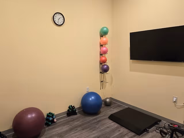 A small exercise room with a wall clock, a mounted flat-screen TV, various exercise balls in different colors and sizes, a set of dumbbells, and a black exercise step platform on a wood floor with beige walls.