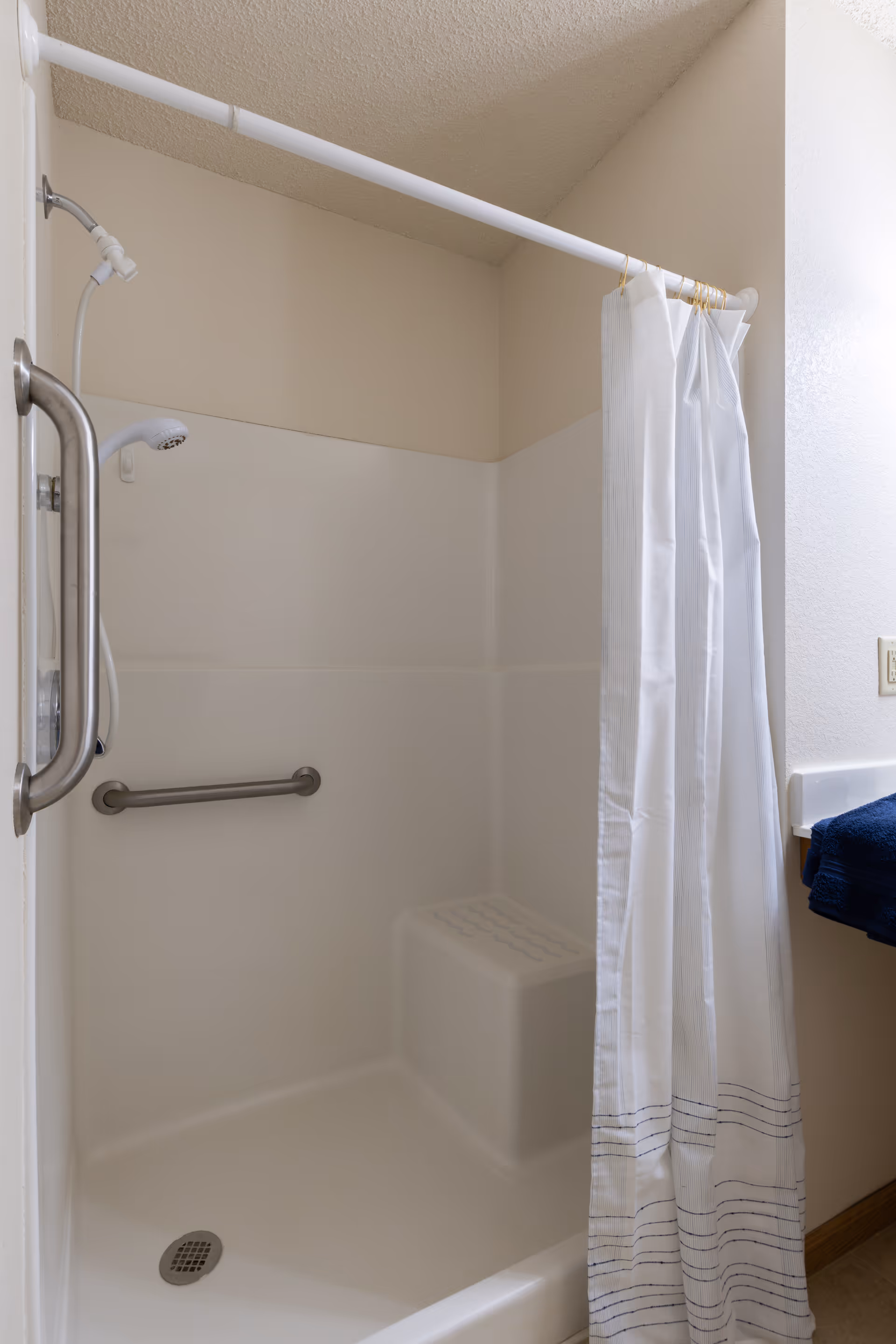 A clean, accessible shower stall with a white curtain partially drawn to the side. The shower features a built-in seat, a grab bar on the wall, and a handheld showerhead. A blue towel is visible on the counter next to the shower.