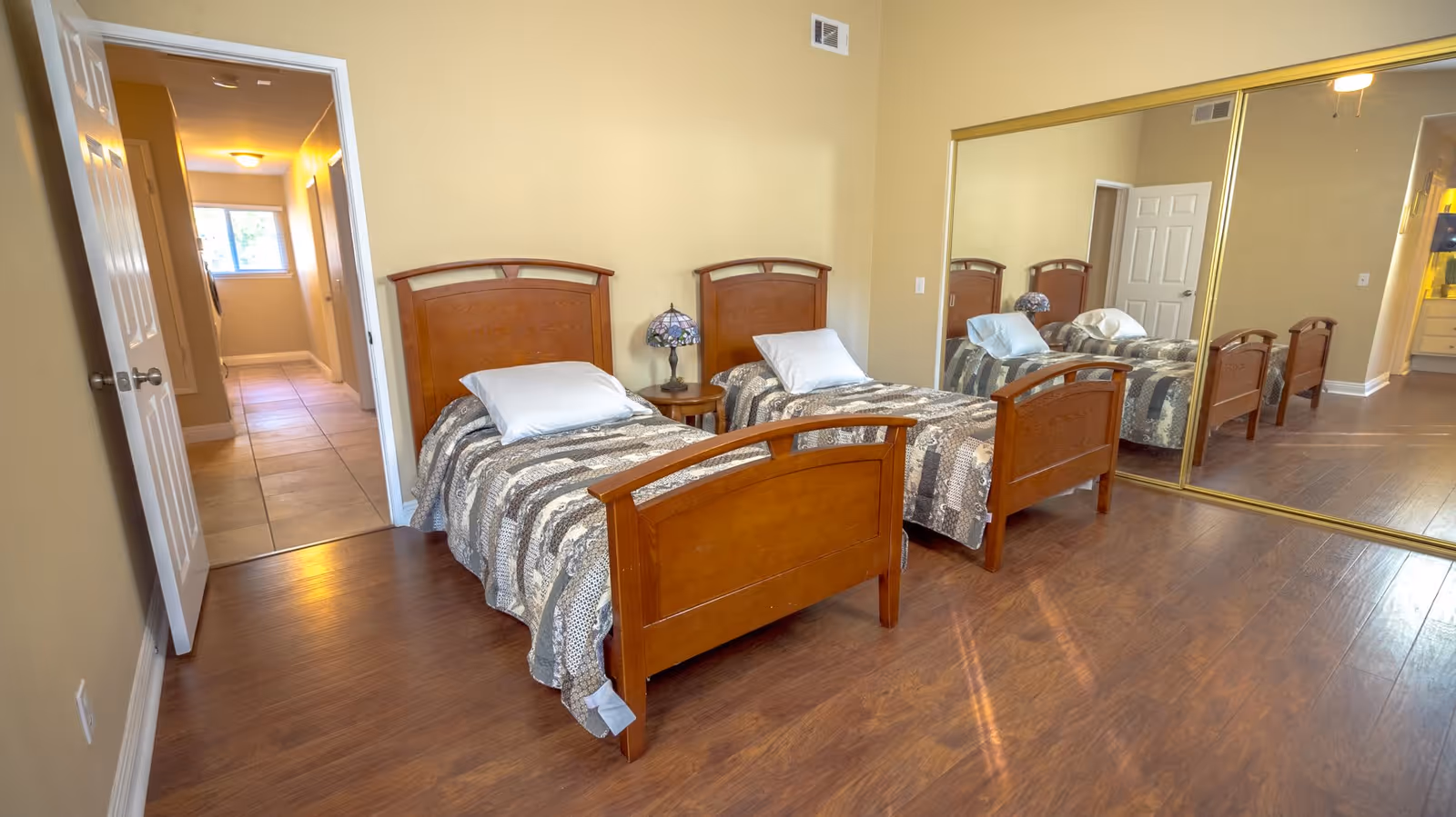 Two twin beds with wooden frames in a bright bedroom with a mirrored closet and hardwood floors.