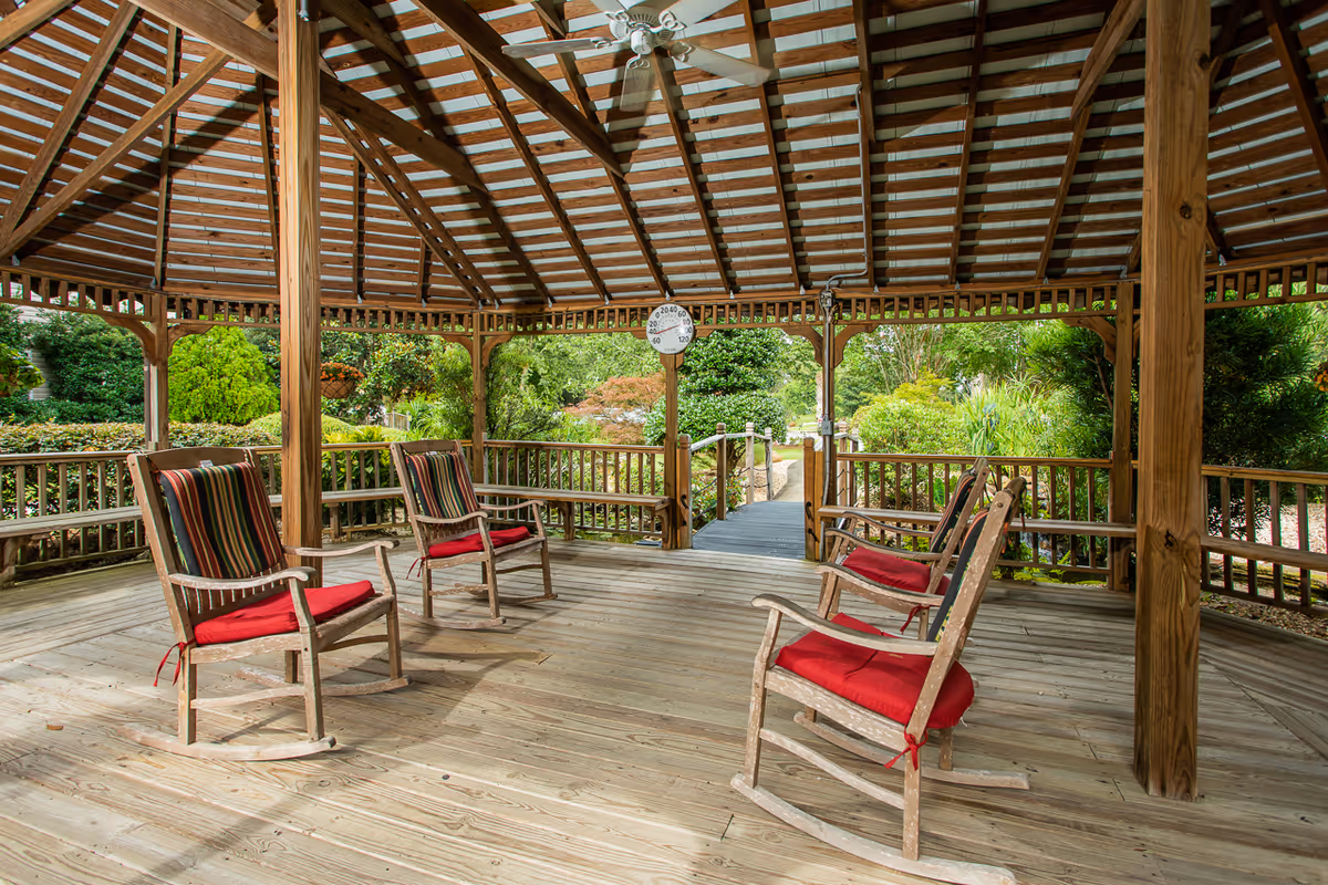 Wooden covered gazebo with several rocking chairs with red cushions overlooking a landscaped garden.
