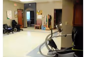 Interior view of a rehabilitation center room with exercise equipment including a stationary bike in the foreground. The room has beige walls and light-colored flooring. There are wheelchairs and chairs along the walls, a bulletin board, and an open doorway leading to another room with a window.