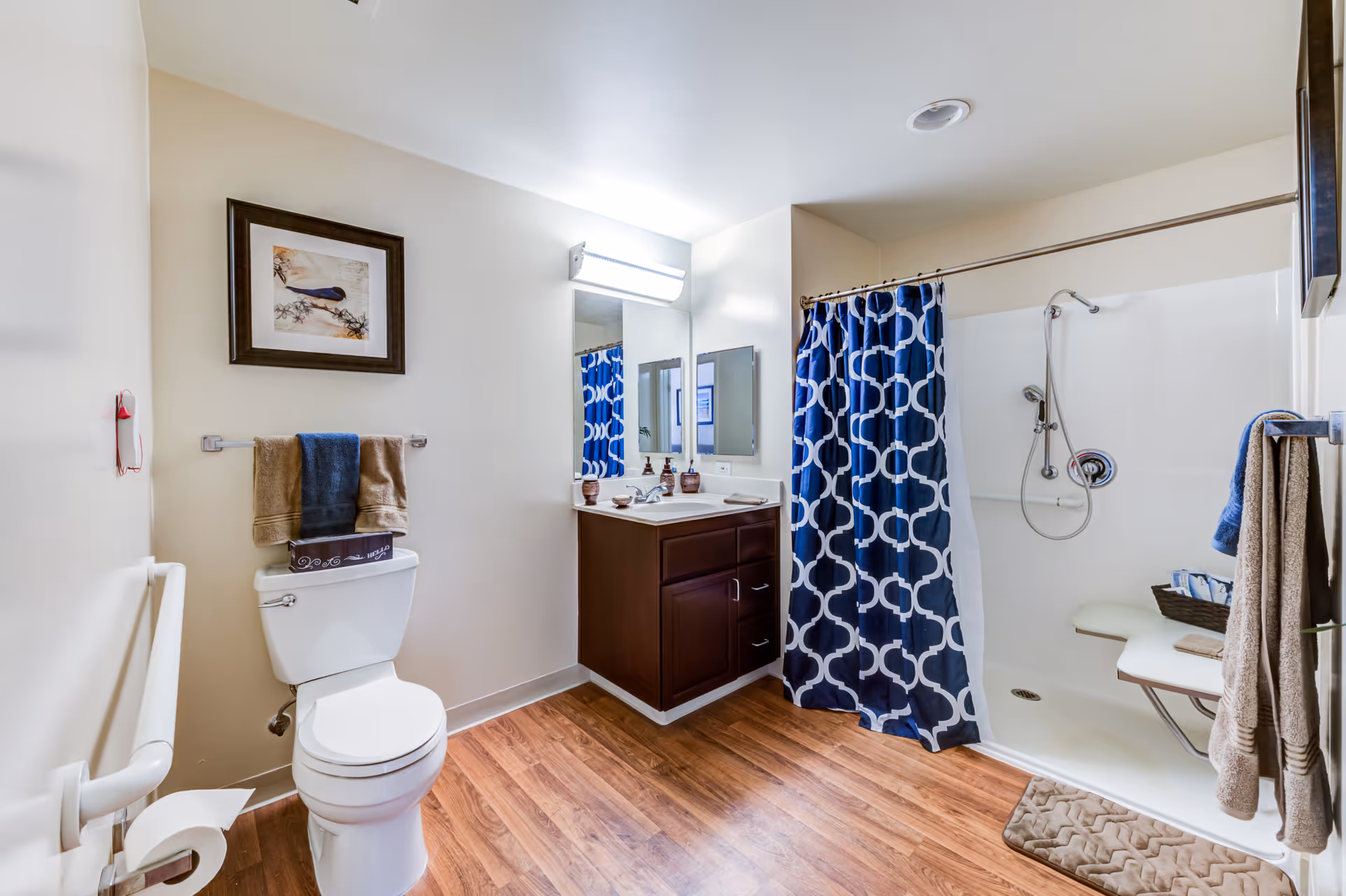 A clean and well-lit bathroom with wood flooring, a white toilet with towels hanging above it, a dark wood vanity with a sink and mirror, and a shower area with a blue and white patterned curtain and a fold-down seat inside the shower.