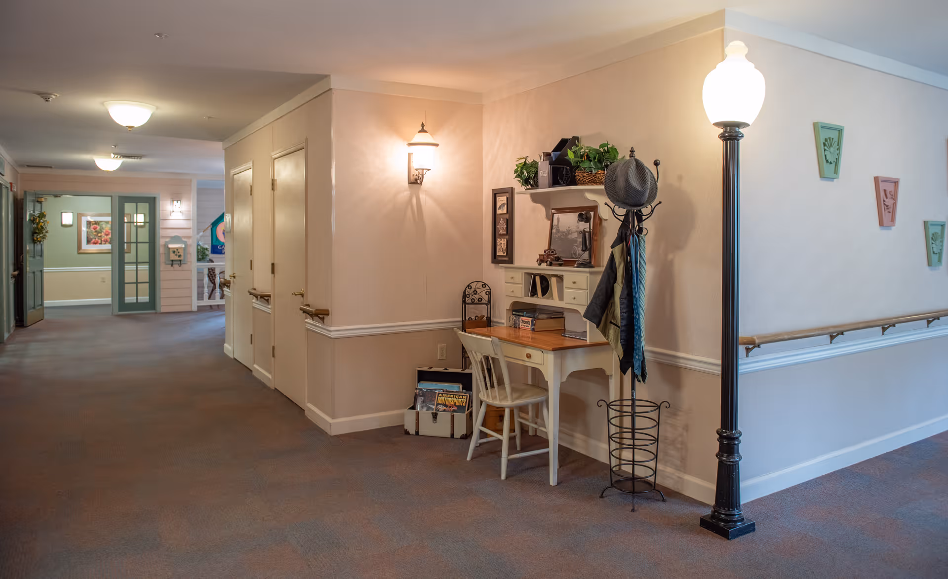 Interior hallway of a senior living facility with beige walls and carpeted floor. On the right side, there is a small white desk with a chair, decorated with plants, books, and framed pictures. Next to the desk is a coat rack holding a hat and some scarves. A tall black lamp post with a glowing white light stands nearby. The hallway extends into the distance with doors and wall decorations visible.