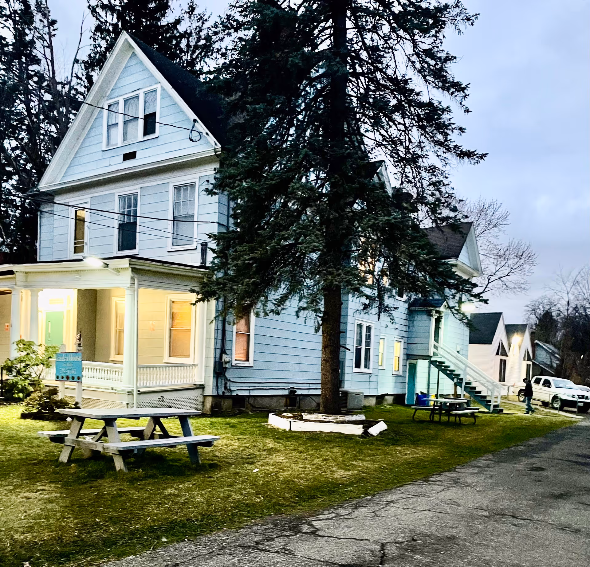 Light-blue multi-story house with a front porch, large evergreen tree, and picnic tables on the lawn at dusk.