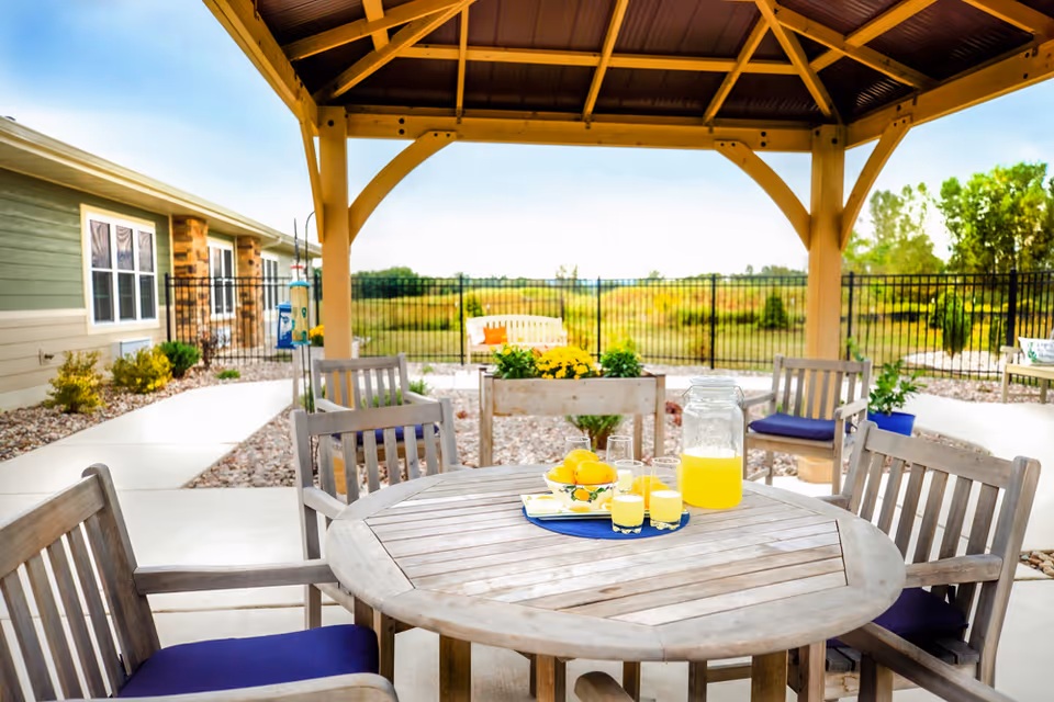 Outdoor seating area under a wooden gazebo with a round wooden table and chairs with blue cushions. On the table, there is a pitcher of lemonade, three glasses filled with lemonade, and a bowl of lemons. The area is surrounded by a paved walkway, plants, and a black metal fence with a grassy field and trees in the background.