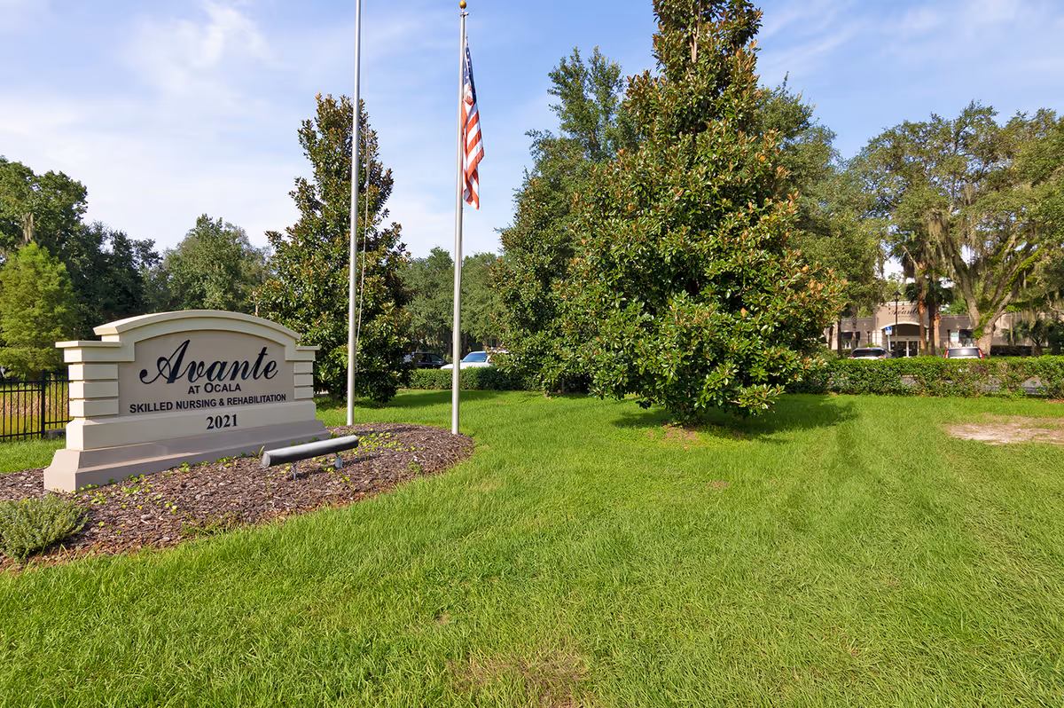 Outdoor view of Avante at Ocala skilled nursing and rehabilitation facility sign on a grassy area with trees and an American flag on a flagpole under a blue sky.
