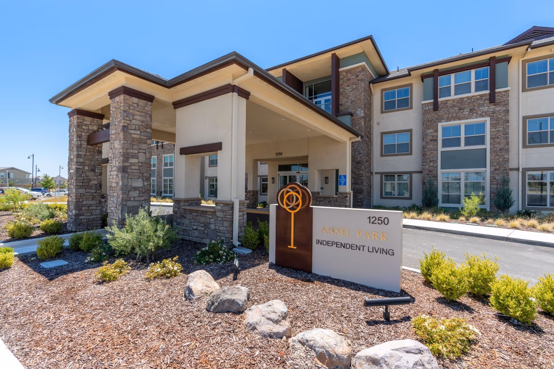 Exterior view of Ansel Park Independent Living facility showing the entrance with stone pillars, a covered drop-off area, landscaped garden with shrubs and rocks, and a sign displaying the facility name and address 1250.