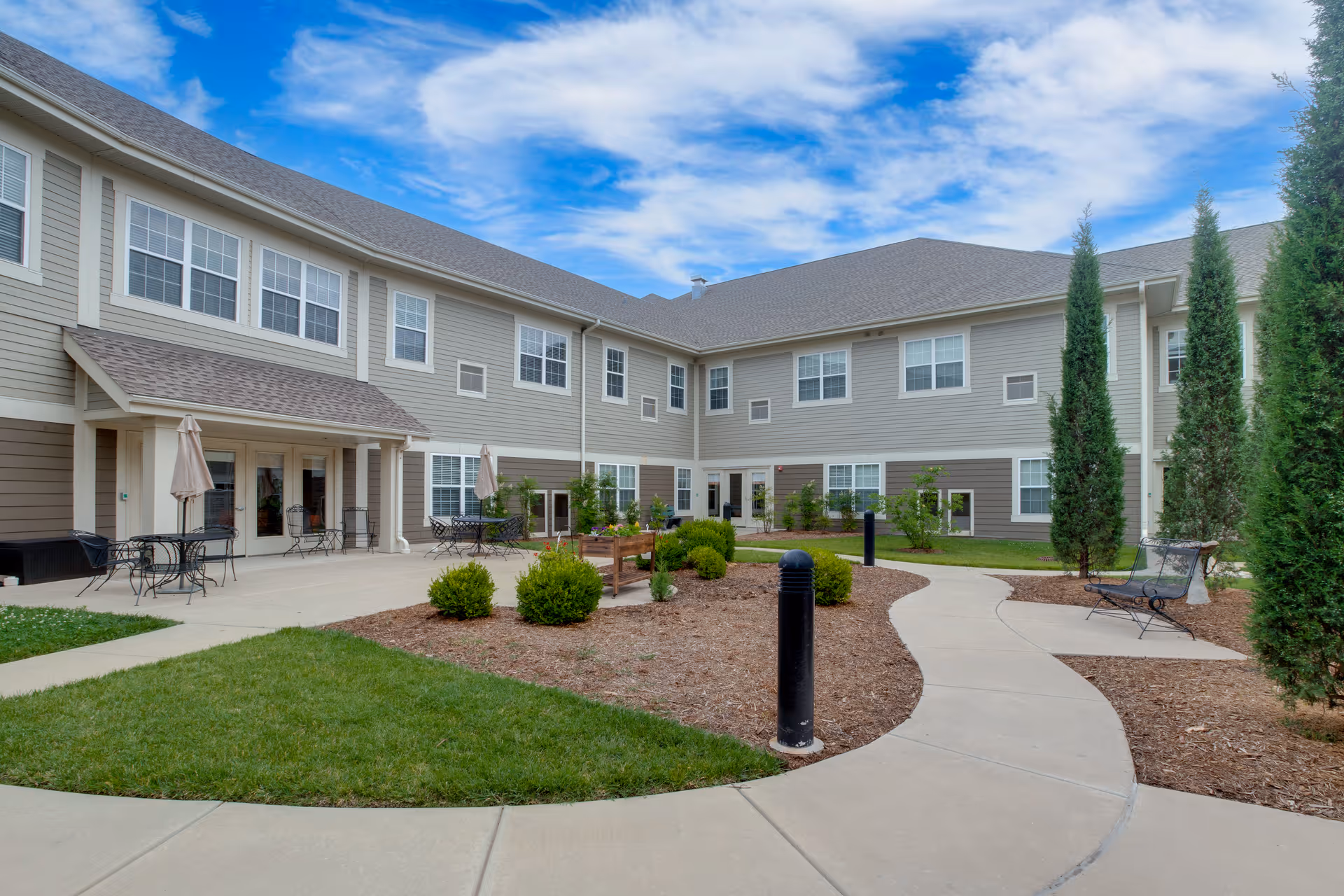 Outdoor courtyard and walkway of a two-story senior living building with patio seating, landscaping, and tall trees.