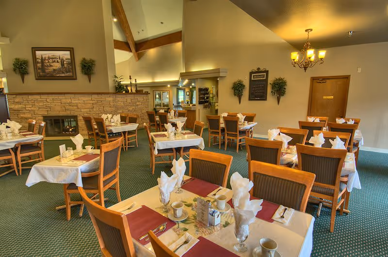 A dining room with multiple tables set for a meal, each table covered with white tablecloths, red placemats, napkins folded in glasses, cups, and utensils. The room has green carpet, wooden chairs, a stone fireplace, wall decorations including plants and a framed picture, and a chandelier hanging from the ceiling.
