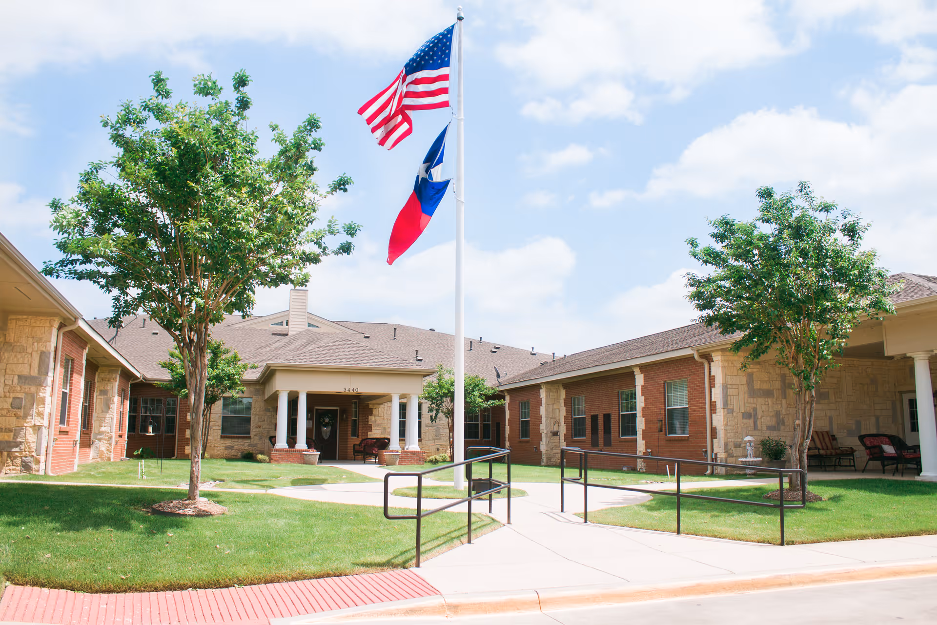 Exterior view of Autumn Oaks of Corinth apartment 2, showing a single-story building with brick and stone facade, a walkway with handrails, green grass, two trees, and flagpoles displaying the American and Texas flags under a partly cloudy sky.