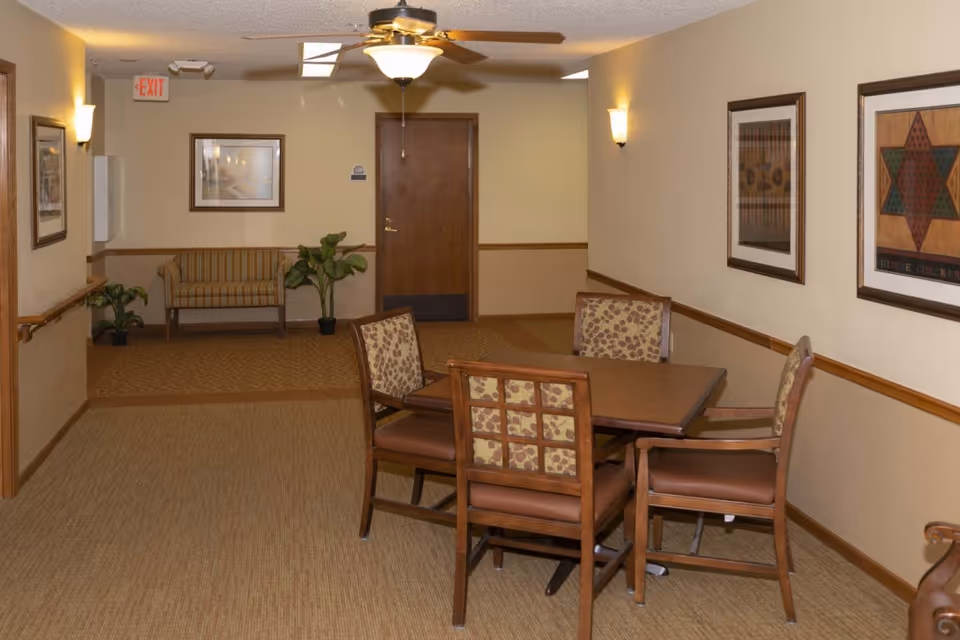 Hallway seating area with a square table and four chairs, framed artwork on beige walls, a bench and potted plants near a closed door.