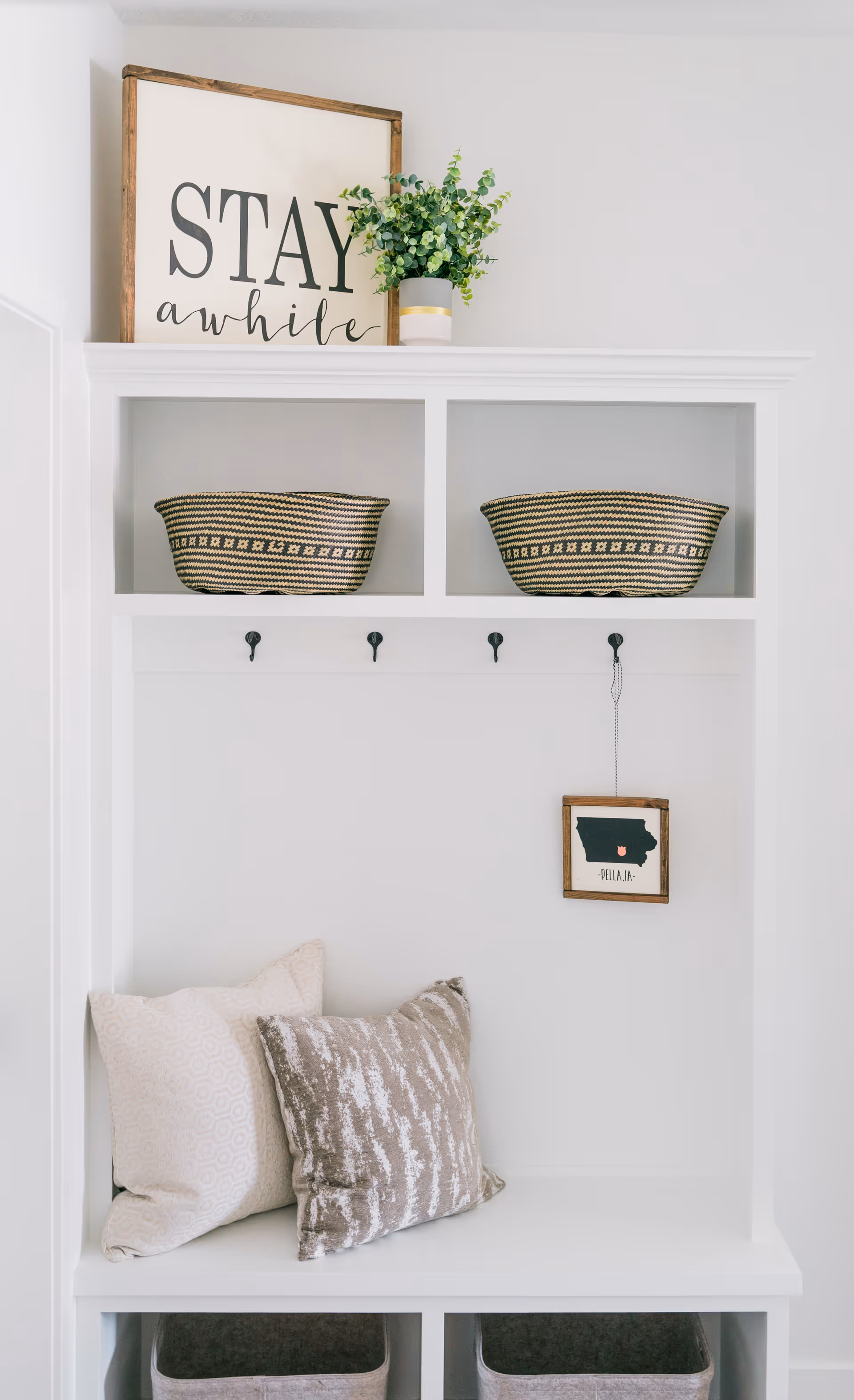White built-in entryway bench with cushions, hooks, wicker baskets, and a framed 'Stay awhile' sign and plant on the top shelf.