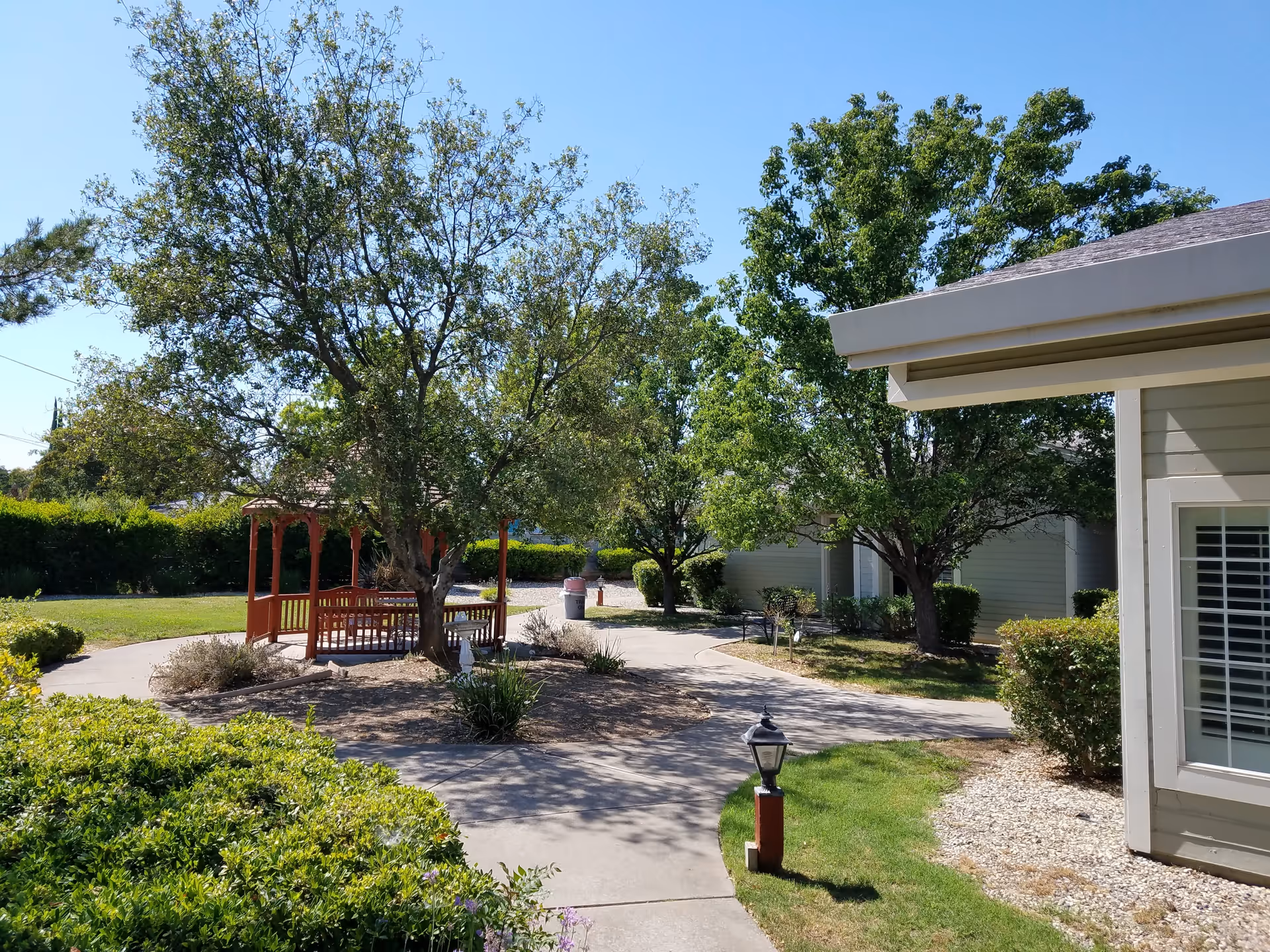 Outdoor garden area at Gramercy Court Assisted Living and Memory Care featuring a paved walkway, green bushes, trees, a red gazebo with benches, and part of a building with a window and white shutters under a clear blue sky.