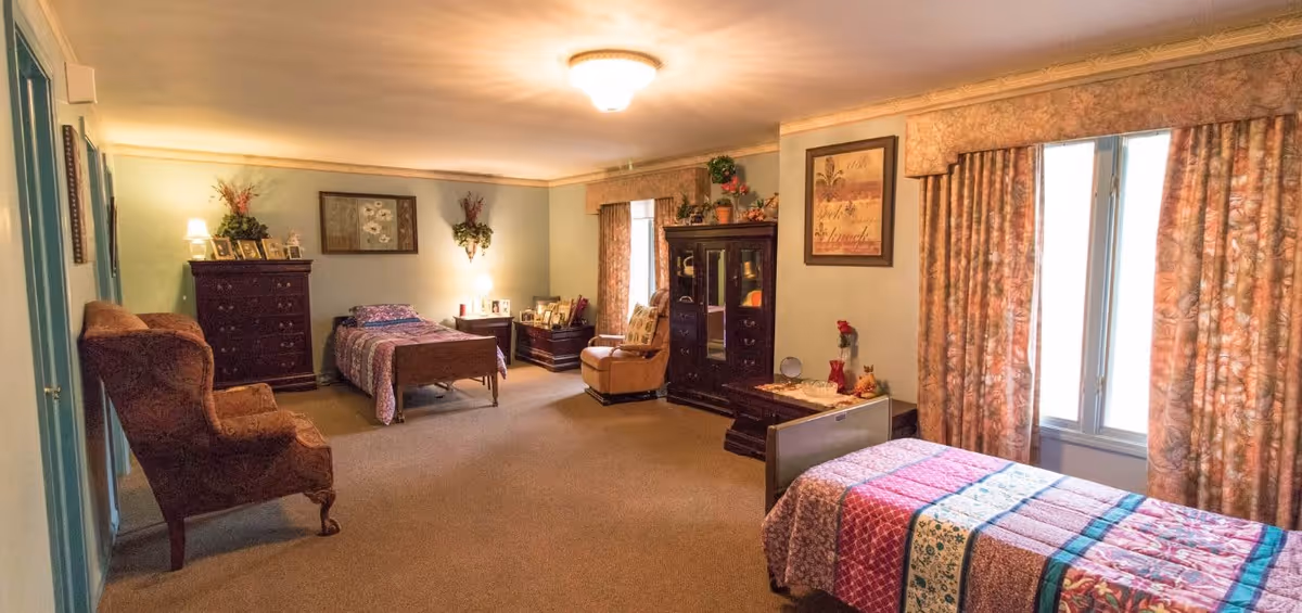 A spacious bedroom in an adult foster care facility with two single beds, each with colorful quilts. The room features a large window with floral curtains, a wooden dresser, a cushioned armchair, and a cabinet with decorative items and framed pictures. The walls are painted light green and decorated with framed artwork and plants.