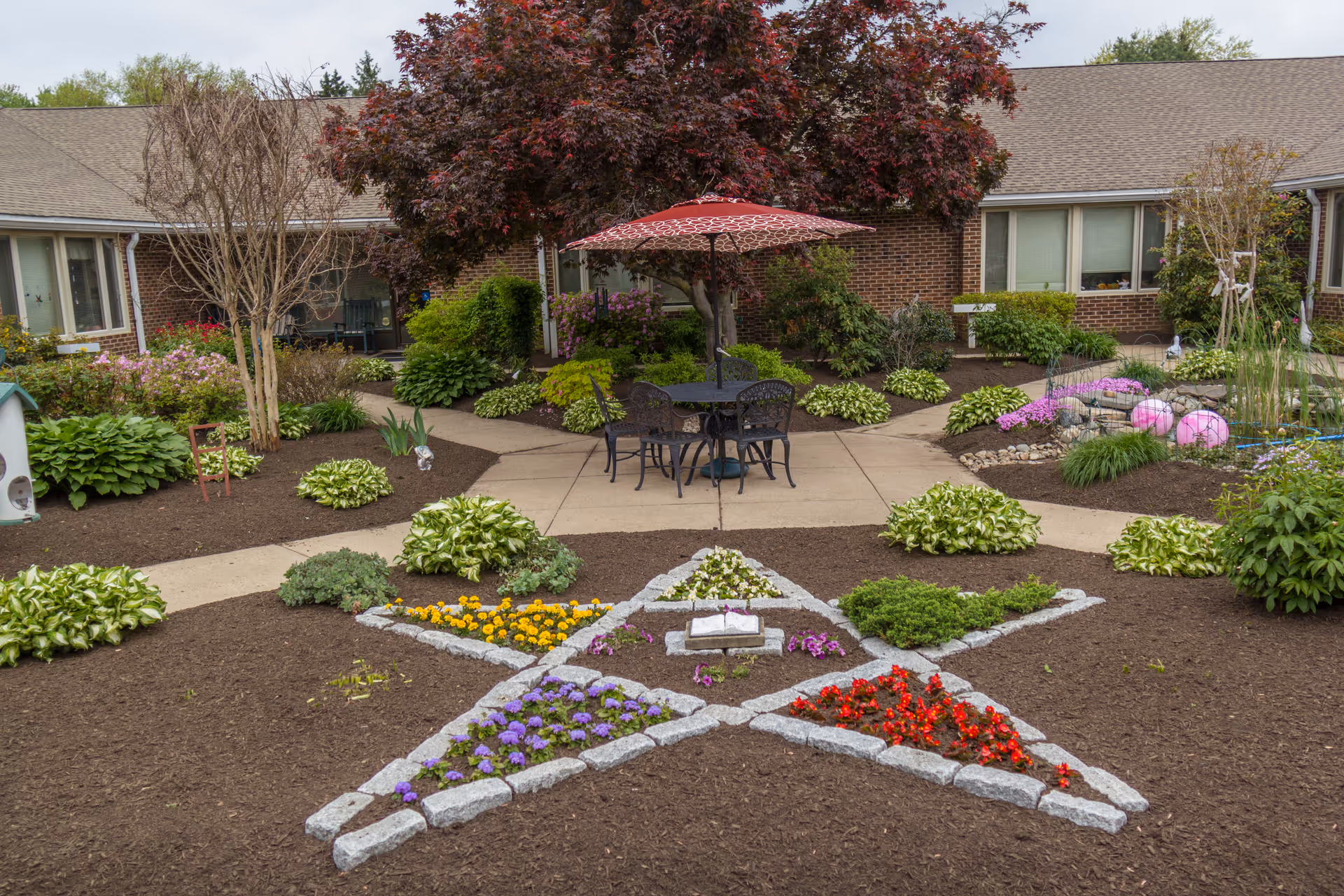 Outdoor garden area at Masonic Village at Warminster featuring a star-shaped flower bed with colorful flowers, surrounded by mulch and various green plants. In the background, there is a brick building with windows and a patio area with a table, chairs, and a red umbrella under a tree with dark red leaves.