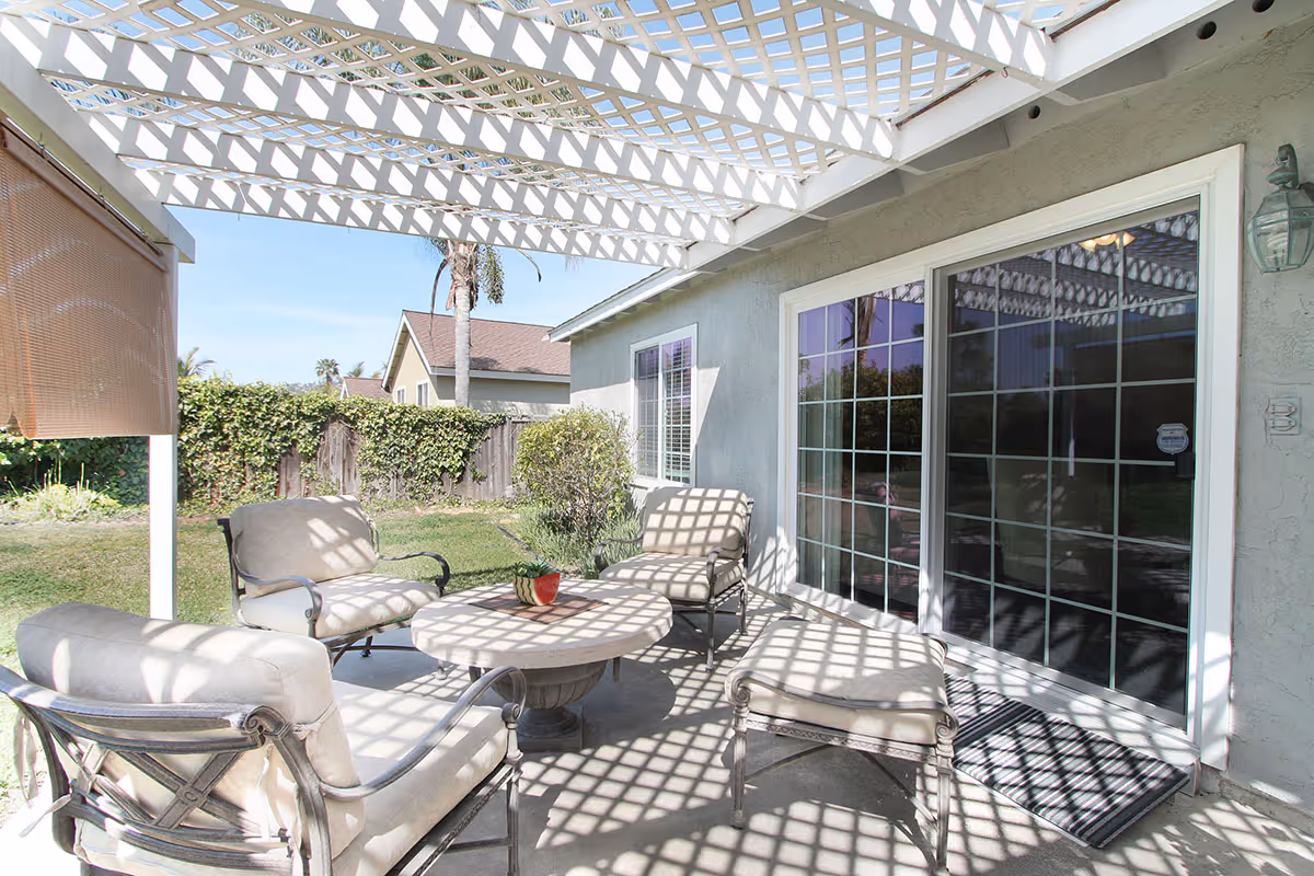 Outdoor patio area with cushioned metal chairs and a round table under a white lattice pergola casting patterned shadows. The patio is adjacent to a house with large sliding glass doors and a small garden with green grass and bushes in the background.