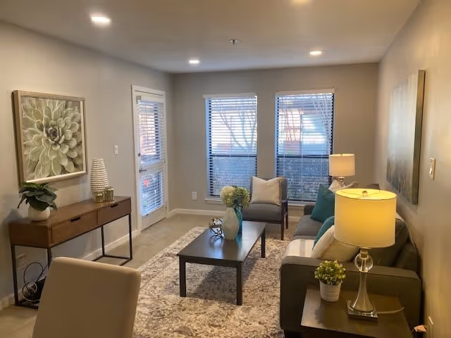 A cozy living room in a retirement community featuring a gray sofa with cushions, a wooden coffee table with decorative items, two armchairs, a side table with a lamp and plant, a console table with a plant and decorative objects, two large windows with blinds, and neutral-colored walls adorned with artwork.