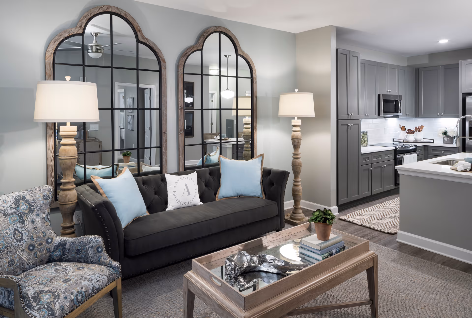 Open-concept living room and kitchen featuring a dark tufted sofa with pillows, patterned armchair, large arch mirrors, wooden coffee table, floor lamps, and grey kitchen cabinets.