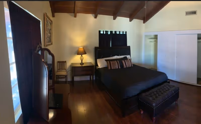 A dark-wood bedroom with a bed, bench, dresser, nightstand, lamp, and vaulted ceiling.