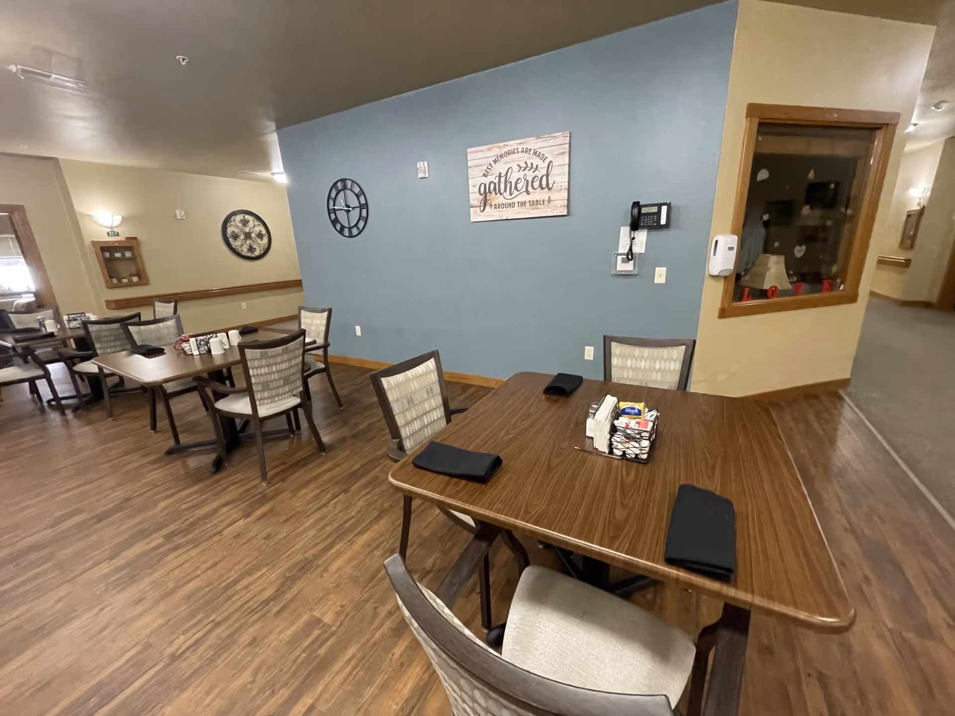 Communal dining room with several wooden tables and upholstered chairs, place settings, and wall decor on a blue accent wall.