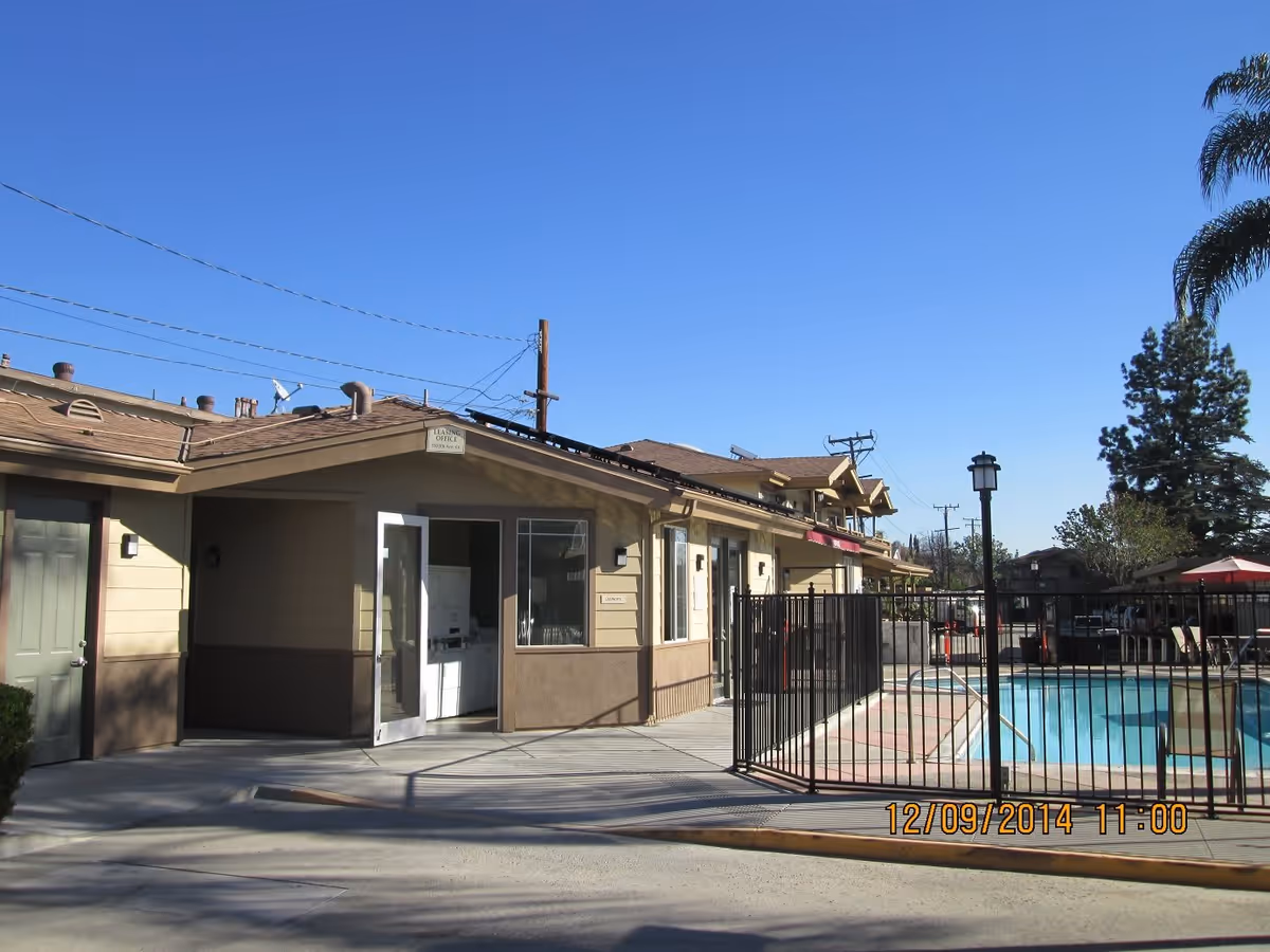 Exterior view of a senior housing building with an open door and a fenced swimming pool under a clear blue sky.