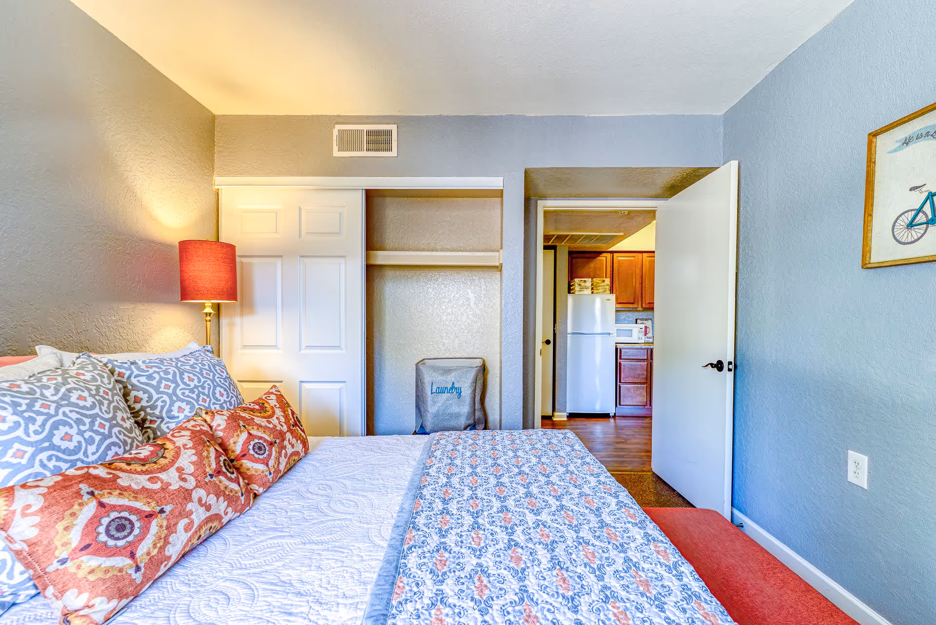 A bedroom with a bed covered in patterned bedding and multiple pillows. To the left of the bed is a lamp with a red lampshade. There is an open closet with sliding doors and a laundry hamper inside. The bedroom door is open, revealing a kitchen area with a refrigerator, microwave, and wooden cabinets. The walls are painted light blue and gray, and there is a framed picture of a bicycle on the right wall.