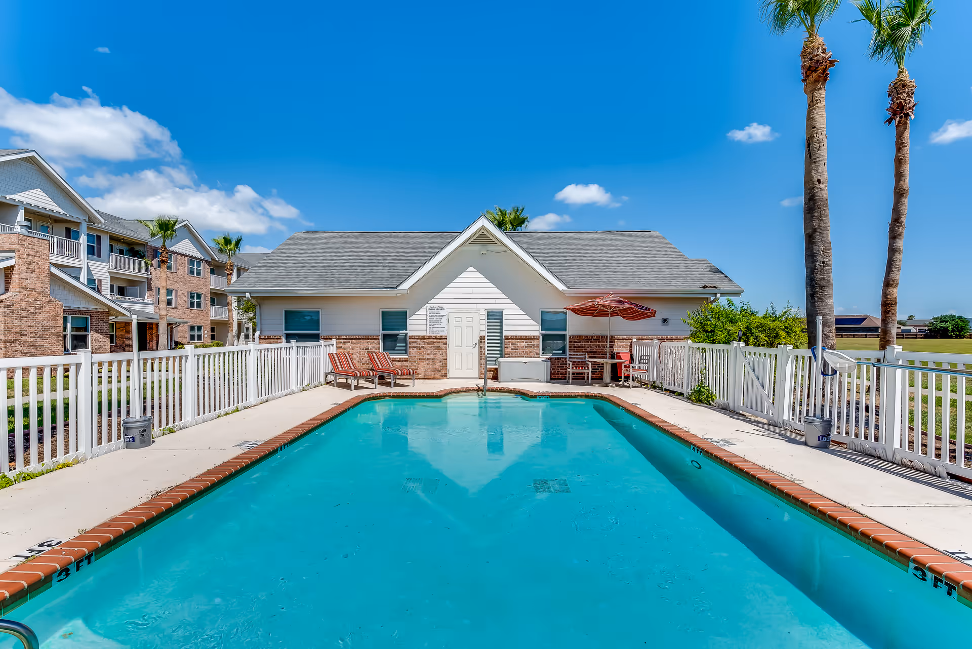 Outdoor swimming pool with clear blue water surrounded by a white fence. There are two lounge chairs with red and white striped cushions on the left side near a small building with a gray roof and white walls with brick accents. On the right side, there is a red umbrella shading a seating area with chairs and a table. Two tall palm trees are visible on the right side, and a multi-story residential building is seen in the background under a bright blue sky with a few clouds.