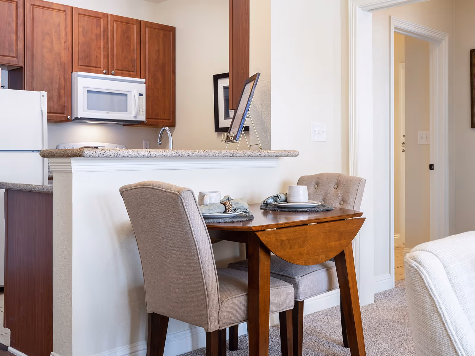 A small dining area with a wooden table set for two with plates, cups, and napkins. Two upholstered chairs are placed at the table. Behind the dining area is a kitchen with wooden cabinets, a white microwave, and a white refrigerator. The room has light-colored walls and carpeted flooring.