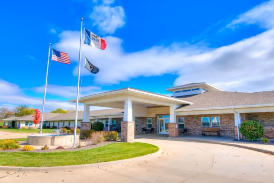 Front entrance of a single-story senior living facility with three flagpoles, a covered porte-cochère, and landscaped grounds.