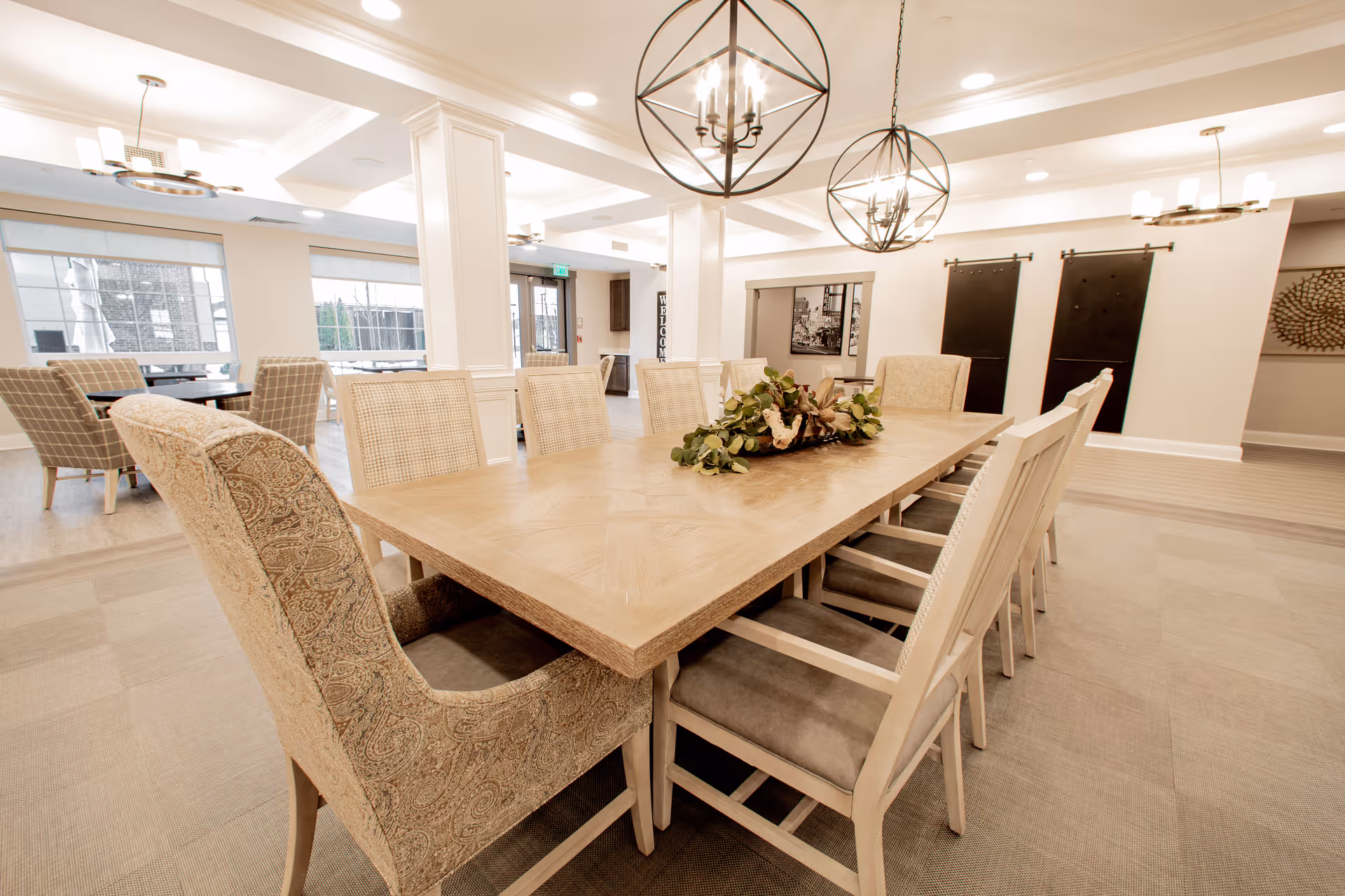 Spacious, well-lit communal dining room with a long wooden table surrounded by upholstered chairs and decorative hanging lights.