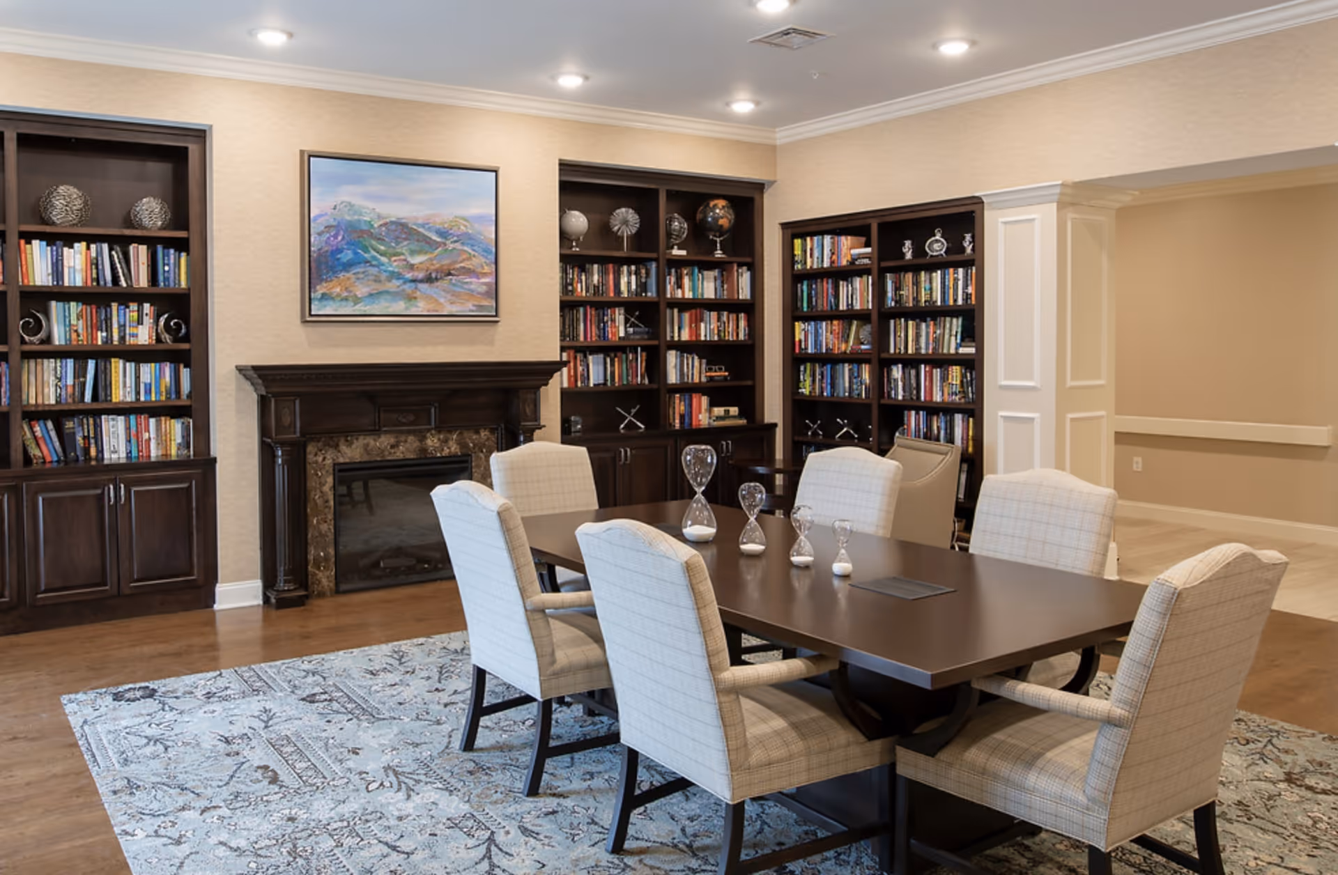 Well-lit community room with a long dark wood table surrounded by upholstered chairs, built-in bookshelves, and a fireplace.