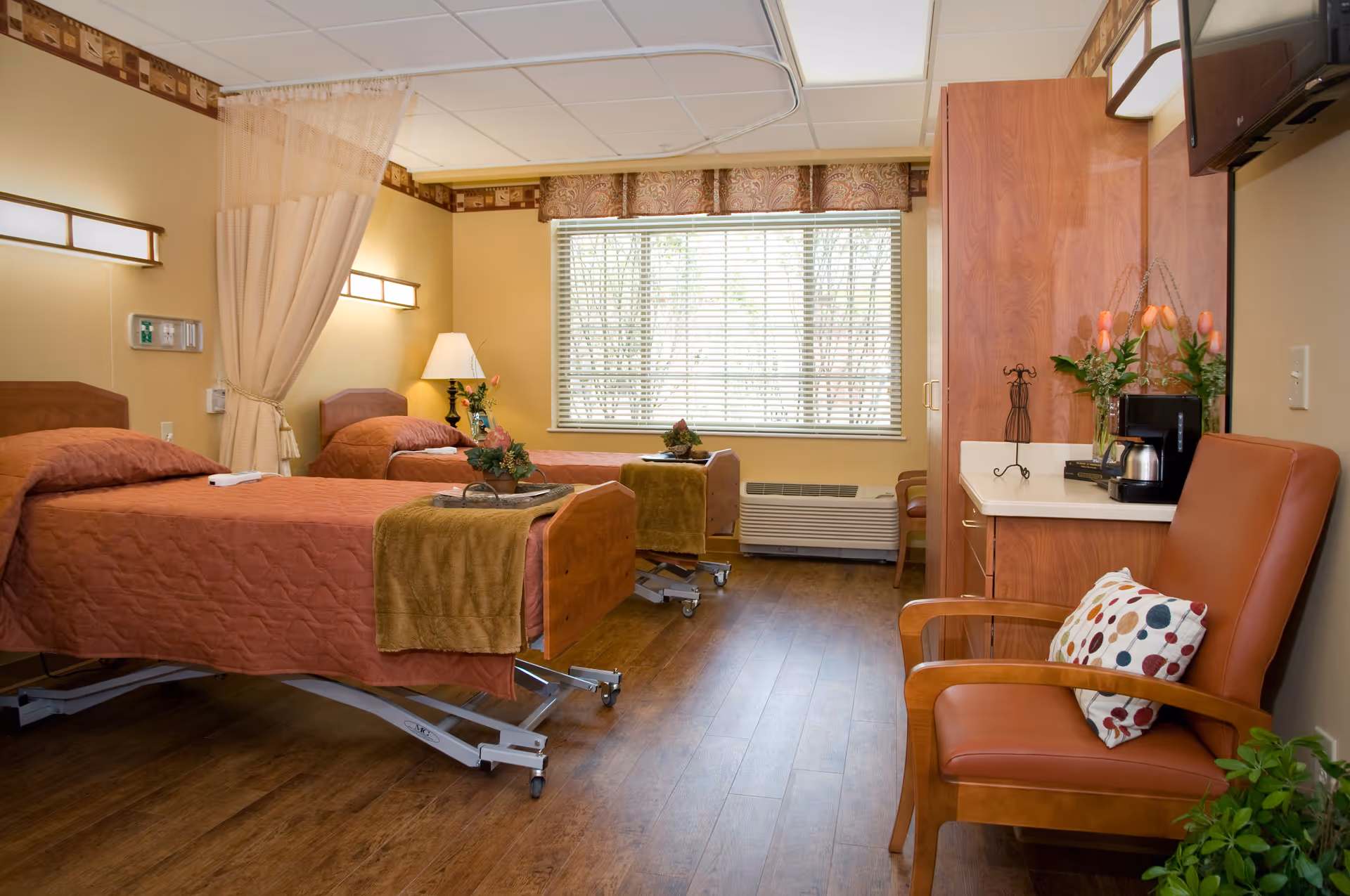 A bright senior care bedroom with two hospital-style beds, wood furniture, a chair, and a window with blinds.
